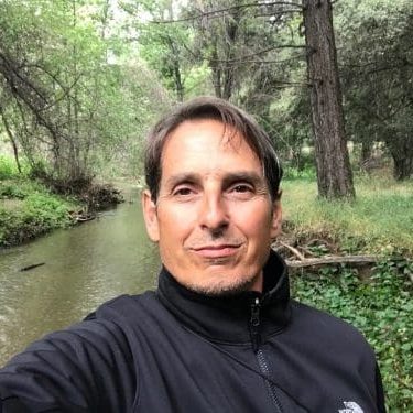 Mark headshot A man wearing a black jacket stands by a narrow creek surrounded by dense trees and greenery, taking a selfie outdoors.